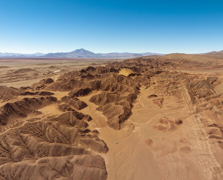 Aerial view of the rugged desert landscape of Tolar Grande featuring deep canyons, undulating sand dunes, and distant mountains under a clear blue sky in Tolar Grande, Salta Province, Argentina.