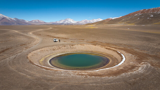 Aerial view of the Balcon del Pissis turquoise lagoon in a vast arid desert landscape, with a vehicle and person nearby, under a clear blue sky in Catamarca, Catamarca Province, Argentina.
