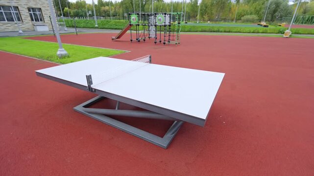 Ping pong table stands on red rubber playground surface. Various climbing frames and slides fill background of modern recreation area