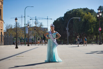 Obraz premium Young woman, brunette, Hispanic, wearing a light blue fantasy dress, diamond crown, and winner's sash of a teen petite beauty pageant, proudly posing in a city square.