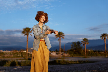 Fototapeta premium Cheerful woman outdoors during golden hour, wearing denim jacket and yellow dress, smiling with relaxed posture, warm light, palm trees and distant mountains in background