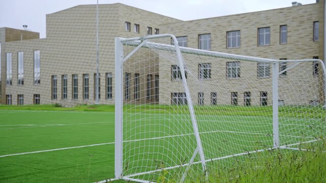 Soccer goal with net stands prominently in foreground of football pitch. Modern tan brick facade of school building visible under cloudy sky