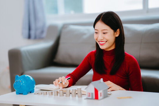Cropped shot of a woman writing in notebook with stacked coins, miniature house, and blue piggy bank on table, property investment and financial planning concept.