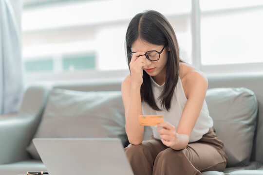 Stressed young Asian woman holding credit card and touching her head while looking at laptop on sofa, financial problems and debt concept.