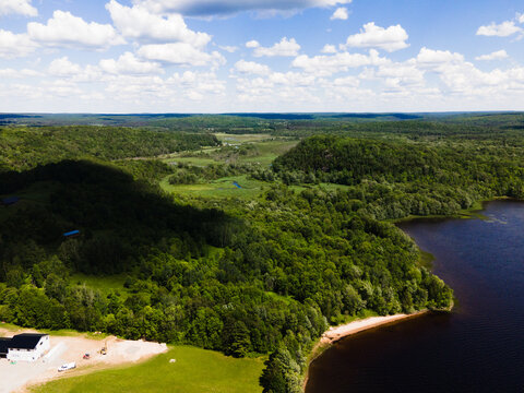 Aerial view of Commanda Lake surrounded by dense forests, rolling hills, and a building near the shoreline under a bright sky in Restoule, Ontario, Canada.