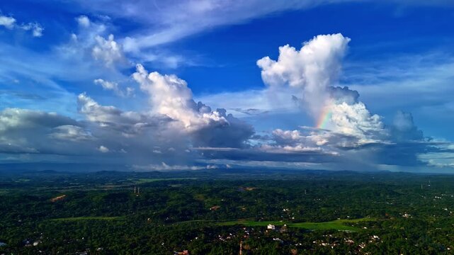 Dramatic rain clouds and a faint rainbow hang over a wide green Sri Lankan landscape under deep blue sky, seen from a drone.