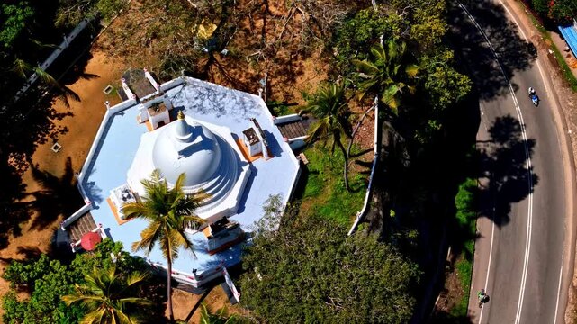 White hilltop stupa sits among palm trees next to a winding roadside as the drone flies past in bright daylight.