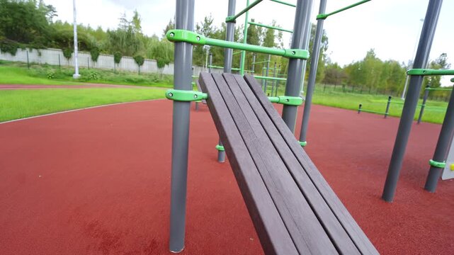 Inclined wooden bench for abdominal exercises sits on rubber surface. Green and grey metal bars complete this outdoor workout station