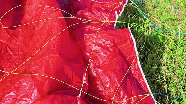 Close up of a red paraglider canopy spread across green grass, with bright suspension lines, wrinkled fabric texture, and outdoor adventure gear ready for a mountain launch in summer.