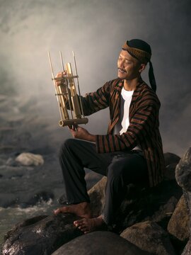 An Indonesian man in traditional Javanese striped lurik shirt and blangkon hat holding an angklung musical instrument while sitting on river rocks.