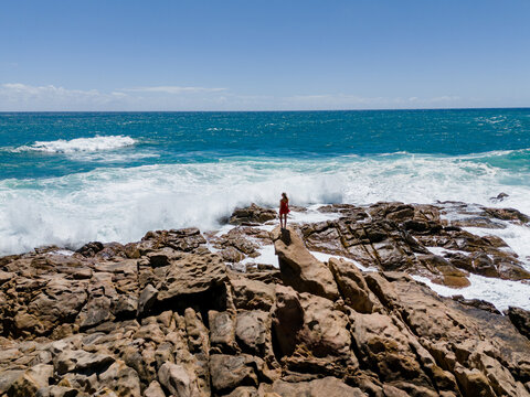 Aerial view of a woman in a red dress standing on the rocky coastline at Injidup Natural Spa with crashing waves and turquoise water in Yallingup, Western Australia, Australia.