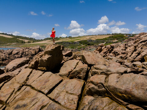 Aerial view of a woman in a red dress standing on the rocky coastline at Injidup Natural Spa, with green hills and blue sky in Yallingup, Western Australia, Australia.