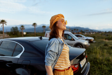 Fototapeta premium A woman enjoys a sunny roadside moment in casual summer style, leaning by a sleek car and gazing toward the horizon. A lifestyle portrait capturing freedom and travel optimism.
