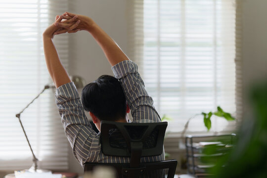 Rear view of Asian man stretching arms at desk in home office for ergonomics and wellness break
