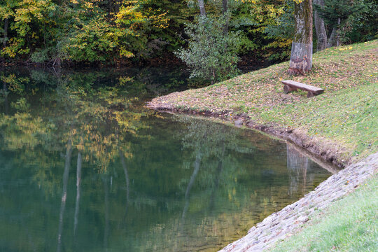 View of the shore of Velka Richnava lake with a wooden bench on a grassy bank surrounded by autumn trees in Stiavnicke Bane, Banska Bystrica Region, Slovakia.