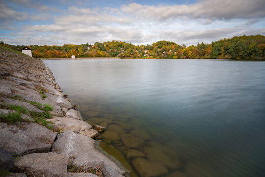 View of Velka Richnava lake with a stone embankment in the foreground and autumn trees under a cloudy sky in Stiavnicke Bane, Banska Bystrica Region, Slovakia.