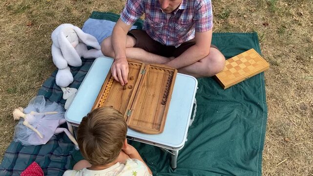 Young father and son spending quality time together in backyard garden, playing board games. Family leisure concept, bonding moments, happiness of simple family life during a summer day outdoors 