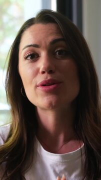 A happy young woman with long brown hair, wearing a white t-shirt, talks to the camera from indoors. She smiles, gestures, and makes eye contact, conveying a friendly and confident demeanor, perfect f