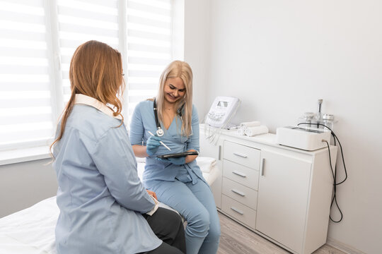 Young woman doctor in blue uniform attentively listens to female patient, reviewing medical records on tablet. Contemporary clinic interior emphasizes trust, care, and advanced healthcare services