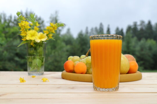 On a wooden desk, on the background of a summer forest landscape, there is a summer still life: a glass of orange juice, wooden tray of fruits and a glass jar of flowers. 