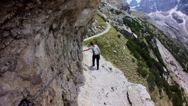 Young woman hiking a narrow, protected path on the side of a mountain, holding onto a safety cable with a deep valley below