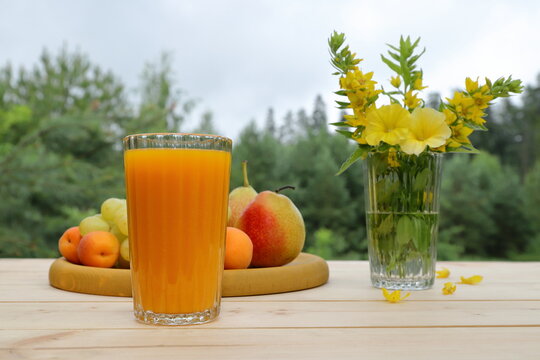 On a wooden desk, on the background of a summer forest landscape, there is a summer still life: a glass of orange juice, wooden tray of fruits and a glass jar of flowers. 