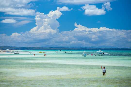 View of Bantayan Island with turquoise shallow waters, traditional outrigger boats, and a woman taking a selfie in the sea in Cebu, Central Visayas, Philippines.