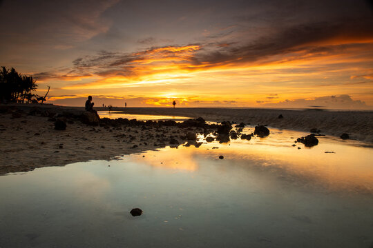 View of Bantayan Island at sunset with a person sitting on a rock by the tide pools, reflecting the golden sky on the sandy beach in Cebu, Central Visayas, Philippines.