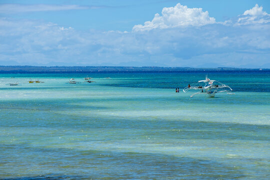 View of traditional outrigger boats floating in the turquoise waters of Bantayan Island with people swimming nearby under a bright blue sky in Cebu, Central Visayas, Philippines.