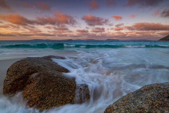 View of Misery Beach with granite boulders, gentle waves washing over the sand, and a colorful sunset sky over the Southern Ocean in Torndirrup, Western Australia, Australia.