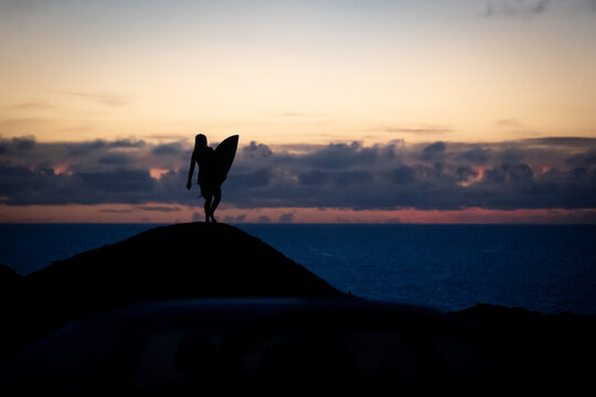 22 January 2022: View of the silhouette of a surfer holding a surfboard on a rocky hill overlooking the ocean during a vibrant sunset