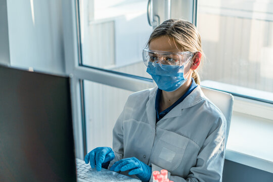 Young woman scientist wearing lab coat, face mask, gloves, and safety goggles sits at desk with computer in bright laboratory. She is conducting scientific research and analyzing experimental results
