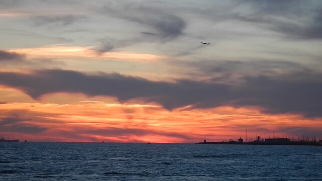 Plane in flight, evening blue sky with orange, red and grey clouds, dark horizon with ship, surface of water on sea, calm small waves and coast in Rome Ostia in Italy. Topics: travel, seascape