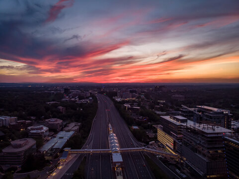 Reston, Virginia, United States - 27 May 2020: Aerial view of the Dulles Toll Road at sunset, featuring a pedestrian bridge, modern office buildings, and a vibrant sky.