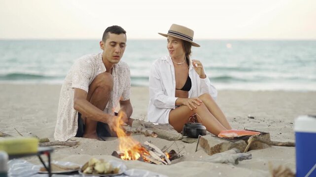 Man and woman spend quality time together on a beach, preparing a meal over a fire with the ocean in the background. Peaceful sunset, summer leisure, and travel lifestyle captured