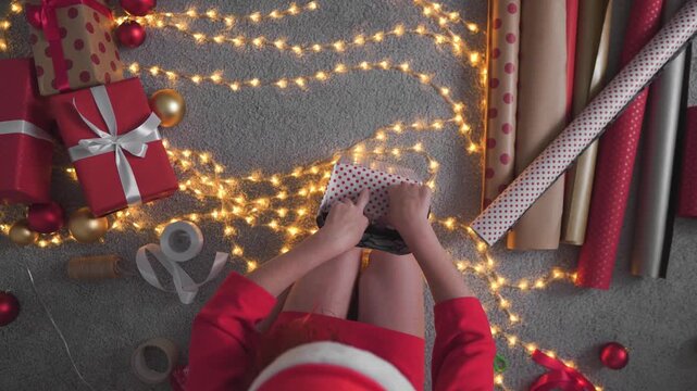 A young child carefully wraps presents amid sparkling string lights, red and gold ornaments, and rolls of wrapping paper. The festive atmosphere captures the joy and anticipation of Christmas