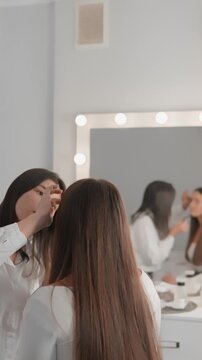 A serene beauty studio scene shows a makeup artist carefully working on a clients eye makeup, reflecting in a bright vanity mirror. The modern interior and soft lighting evoke a calm, trendy vibe