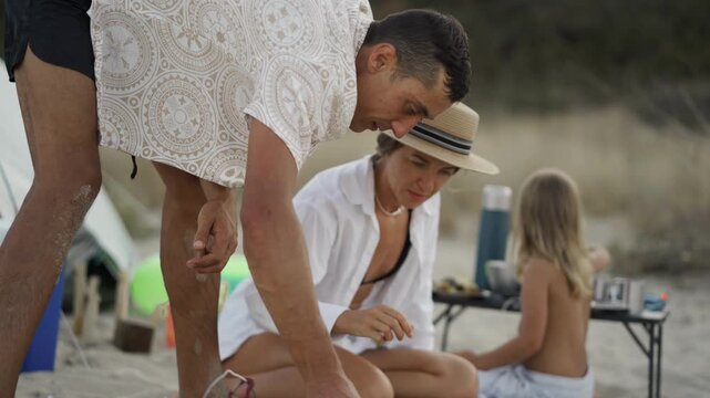 A joyful family scene unfolds as parents and their child relax on the sandy beach, surrounded by nature. Together, they savor snacks and create lasting memories during a sunny summer outing