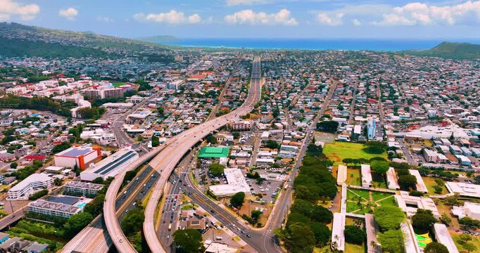 Highway interchange cutting through Honolulu residential district, Hawaii. Multilane freeway converging toward ocean, flanked by low density housing, green hills and mountains backdrop.