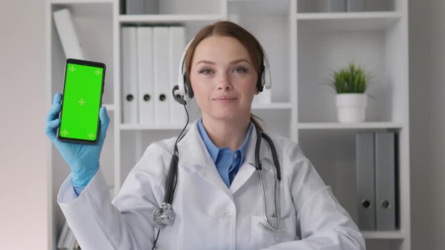 A professional woman doctor in a white coat and stethoscope presents a green screen phone for medical app promotion. She sits in a bright office, ready for online consultation