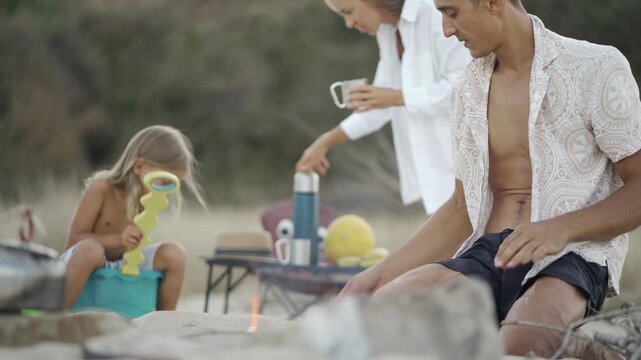 A family spends quality time outdoors, bonding around a campfire in a scenic natural setting. The relaxed mood and casual clothing highlight the joy of summer camping adventures