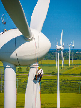 Grobina, Latvia - 14 June 2025: Aerial view of a technician suspended by ropes performing maintenance on a wind turbine at Grobinas veja parks with green fields in the background.