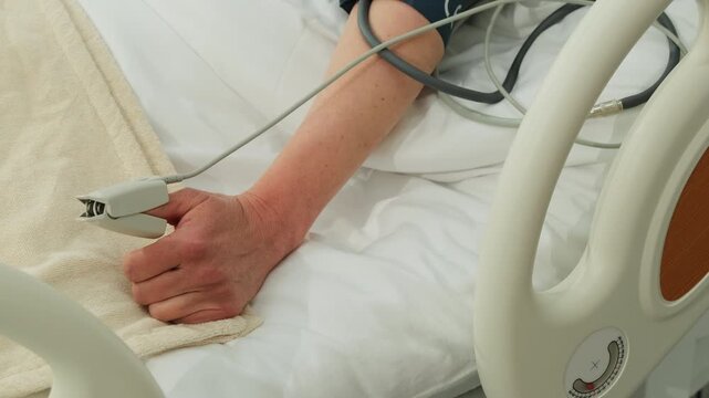 Woman holds pulse oximeter in hand while resting in hospital bed. Sensor records fingertip data tracking patient condition during monitoring