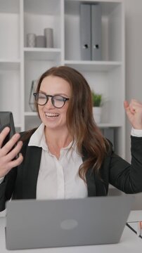 Confident female entrepreneur experiences joyful moment while working remotely at her desk. Happy office worker celebrates achievement using mobile phone and laptop in bright workspace