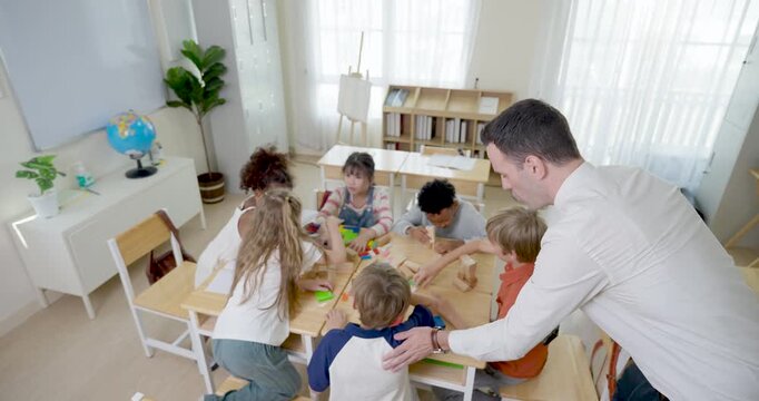 Overhead classroom view shows male teacher guiding multicultural children placing colorful blocks on shared desk, group activity builds teamwork, math thinking, peer support, school engagement