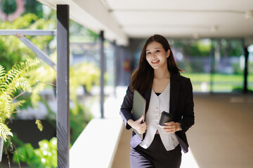 Fototapeta premium Asian professional businesswoman confidently walking outdoor through a modern office building hallway. Carrying a laptop and a coffee cup. Embodying success. Career advancement