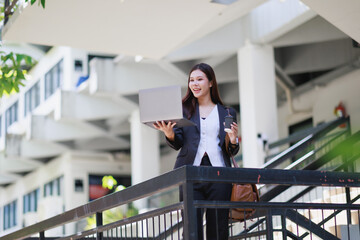 Fototapeta premium Asian businesswoman communicating during a video call on laptop, holding a coffee cup, carrying a work bag, walking down modern office building stairs outside