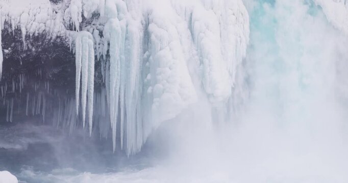 View of a frozen waterfall with large icicles hanging from the rocky cliff face above the icy blue water in Iceland.