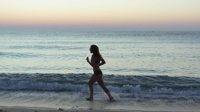 A fit woman runs on the beach at dawn, her silhouette highlighted against the tranquil water and colorful horizon. The serene scene captures wellness, motivation, and coastal lifestyle