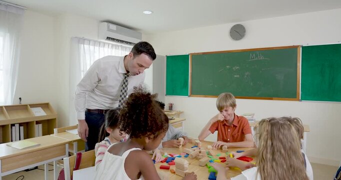 Male primary teacher assists elementary pupil group arranging colorful blocks at shared desk, classroom teamwork lesson builds problem solving, arithmetic readiness, participation, motivation